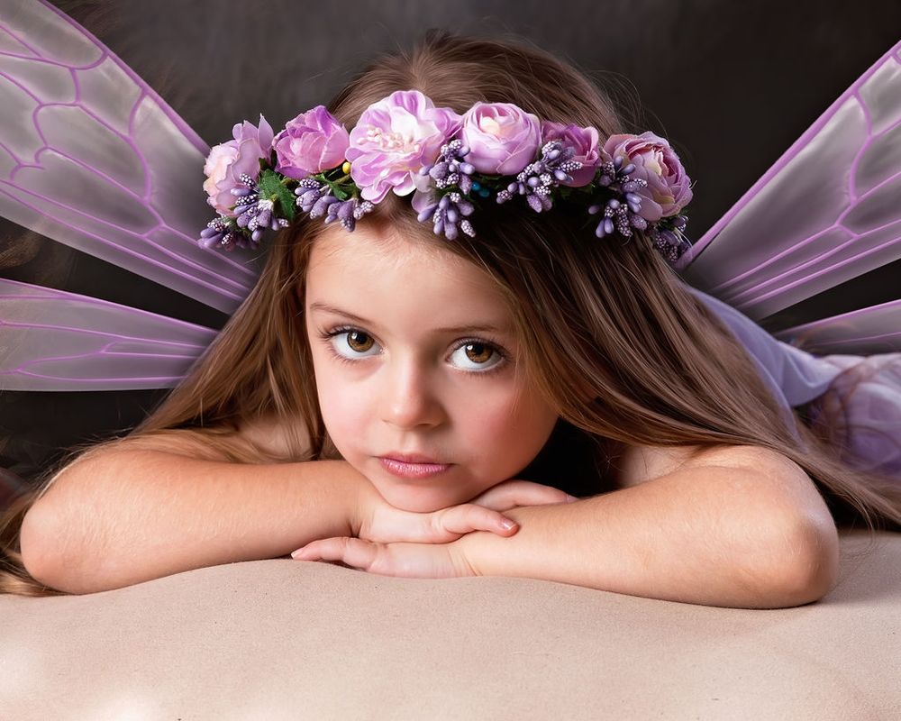 Young girl dressed as a fairy with purple wings and a floral crown of pink and lavender flowers poses gracefully.