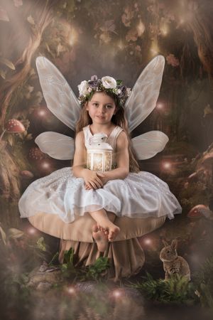 Young girl dressed as a fairy in white dress with wings and flower crown sits on mushroom in enchanted forest.