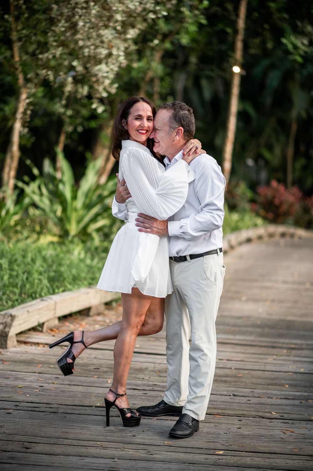 A romantic couple in white embraces on a wooden boardwalk surrounded by lush tropical foliage.