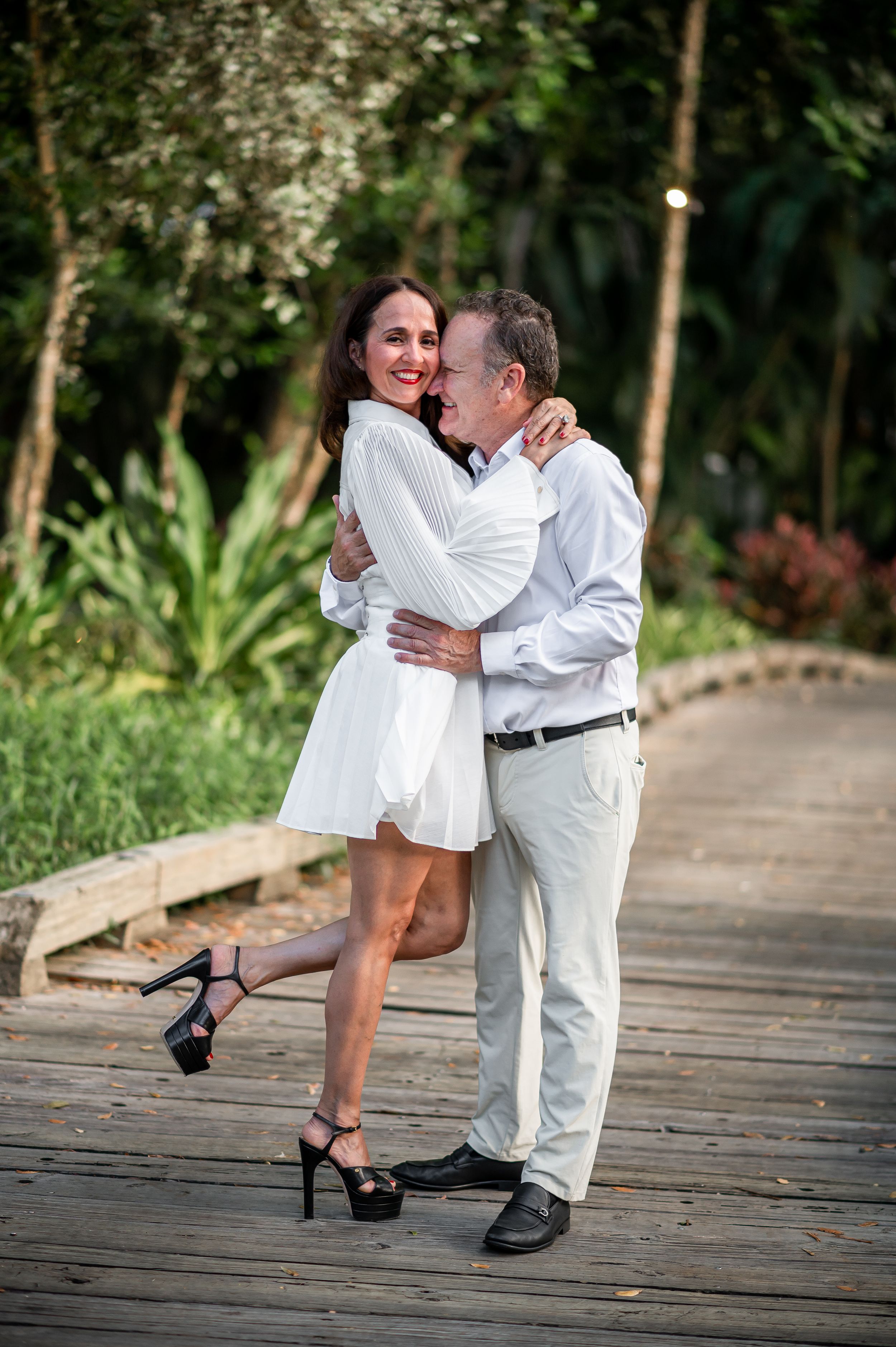 A romantic couple in white embraces on a wooden boardwalk surrounded by lush tropical foliage.