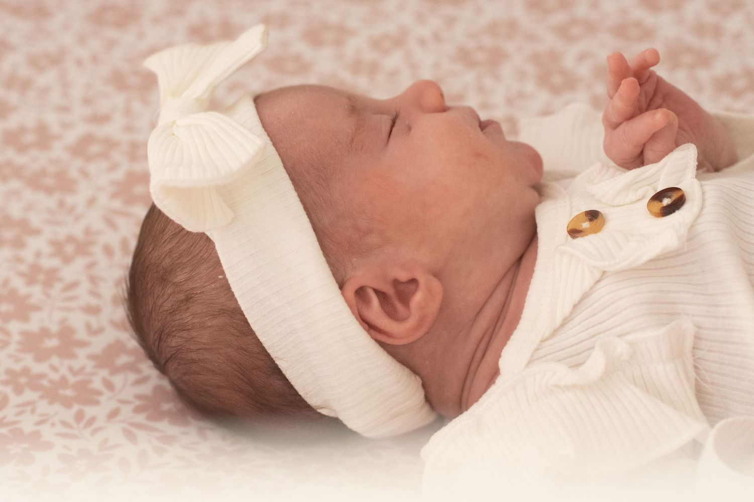 A newborn baby wearing a white knit headband and white outfit laying on textured beige fabric.