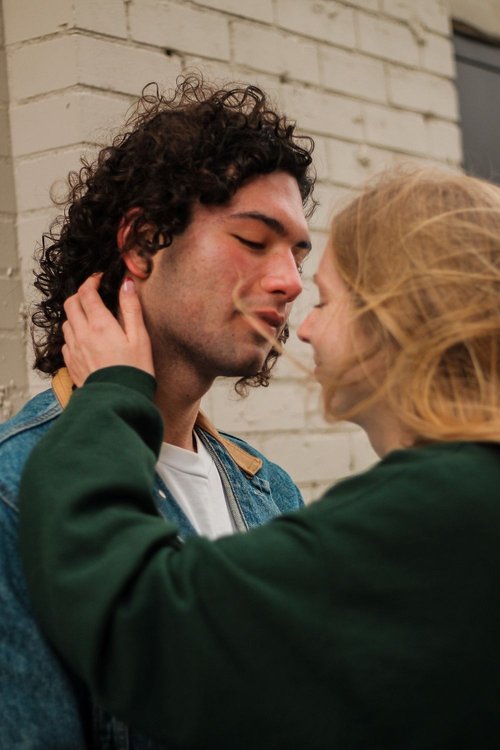 Couples photography with a curly haired man against a wall and the woman leaning in to kiss.