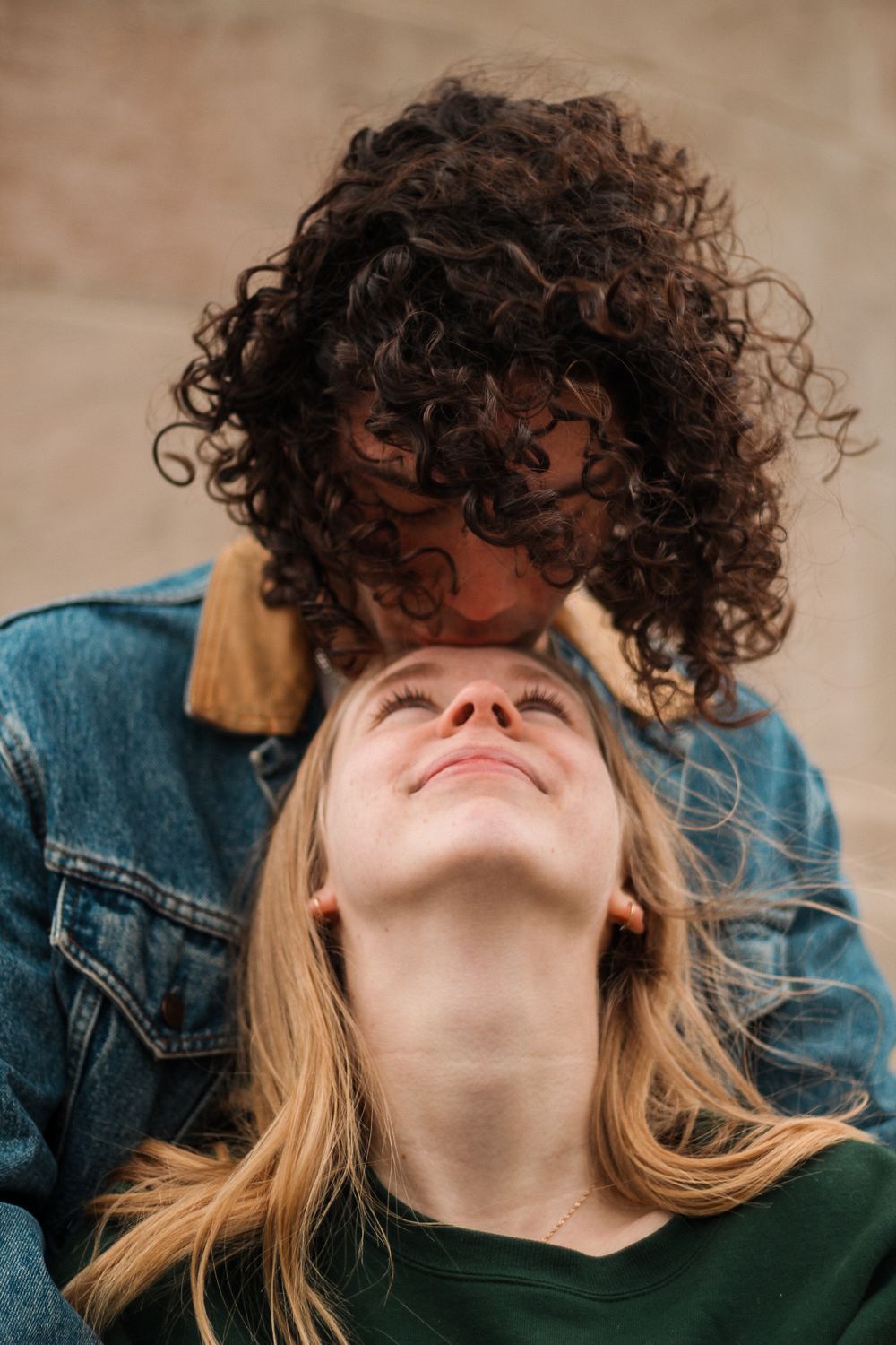Couples photography with a curly haired man behind a blond woman, and looking down while the woman looks up.