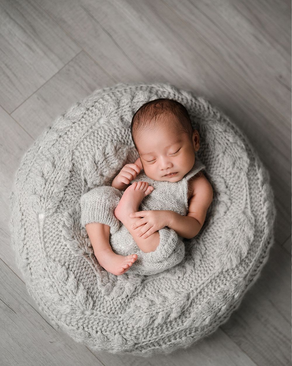 A sleeping newborn baby wrapped in a cozy gray knit blanket on a light wooden floor in a professional photo series.