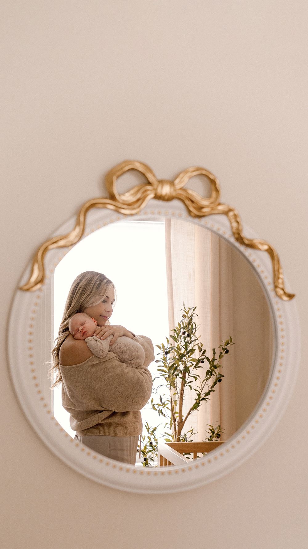 A series of oval mirrors with gold bows reflect a mother with the baby in beige clothing and decorative greenery against a soft pink wall.