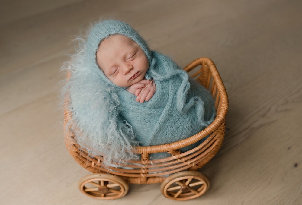 Sleeping newborn wrapped in soft blue blanket nestled in vintage wicker doll carriage for professional photo session.