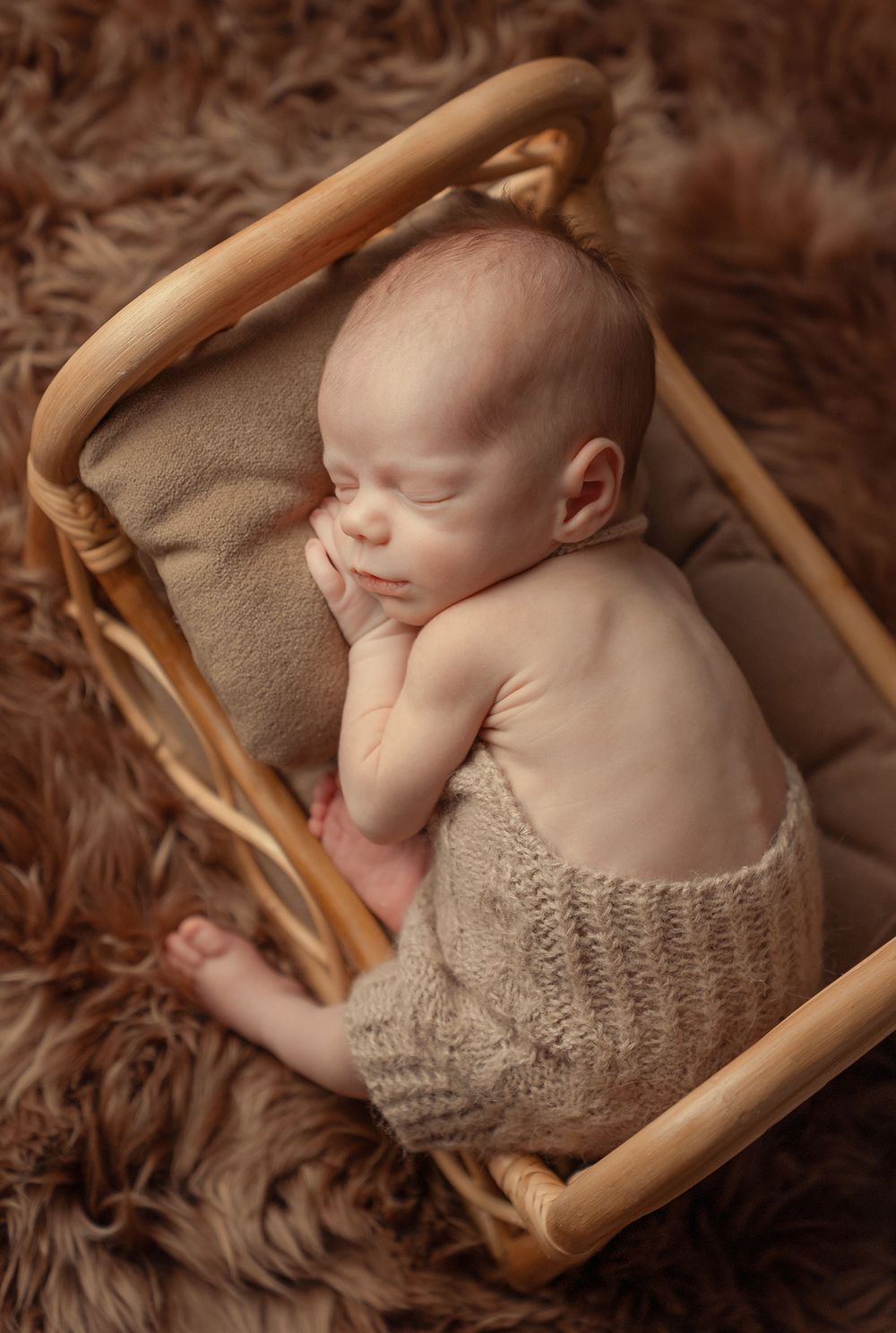 Sleeping newborn rests peacefully in wooden rocking chair with knit wrap and brown fur blanket in warm sepia tones.