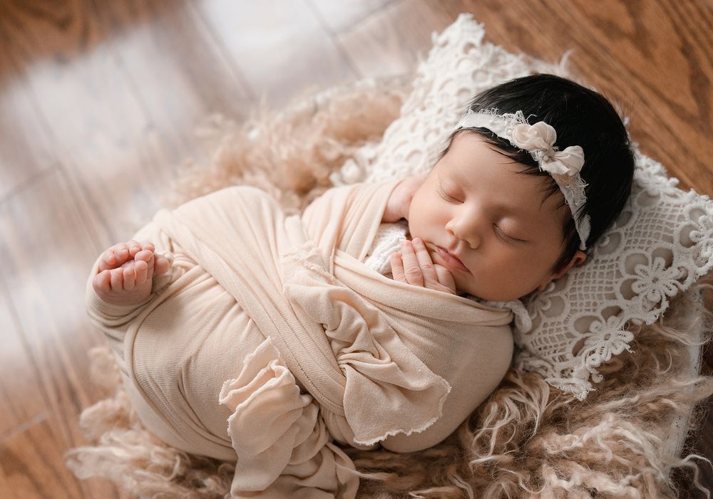 Newborn baby sleeping peacefully on white lace blanket wearing soft peach-colored outfit.