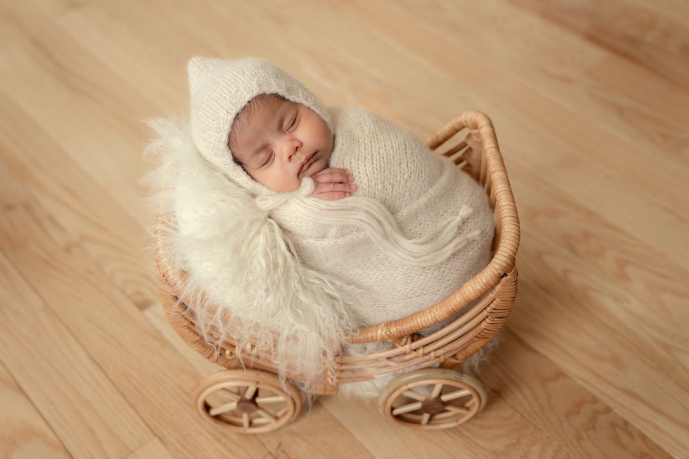 A sleeping newborn wrapped in white swaddle lies in a decorative wicker carriage with fluffy white fur lining on wooden floor.