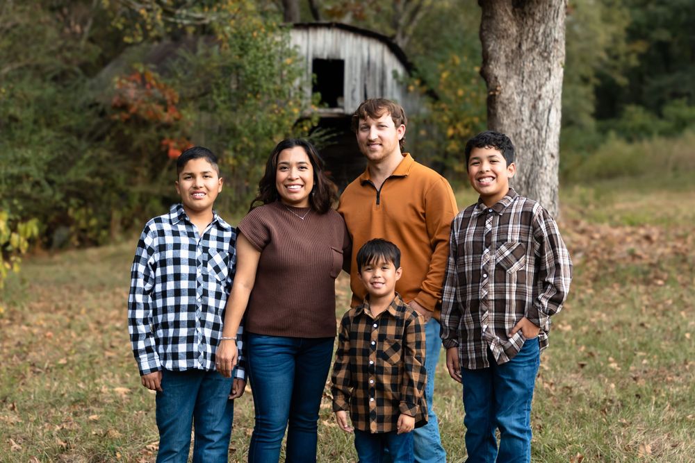 A family stands together outdoors in fall clothing with autumn foliage and a rustic barn in the background.
