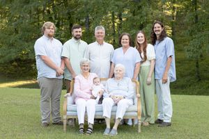 Three generations gather for a family portrait outdoors with a bench and trees in background.