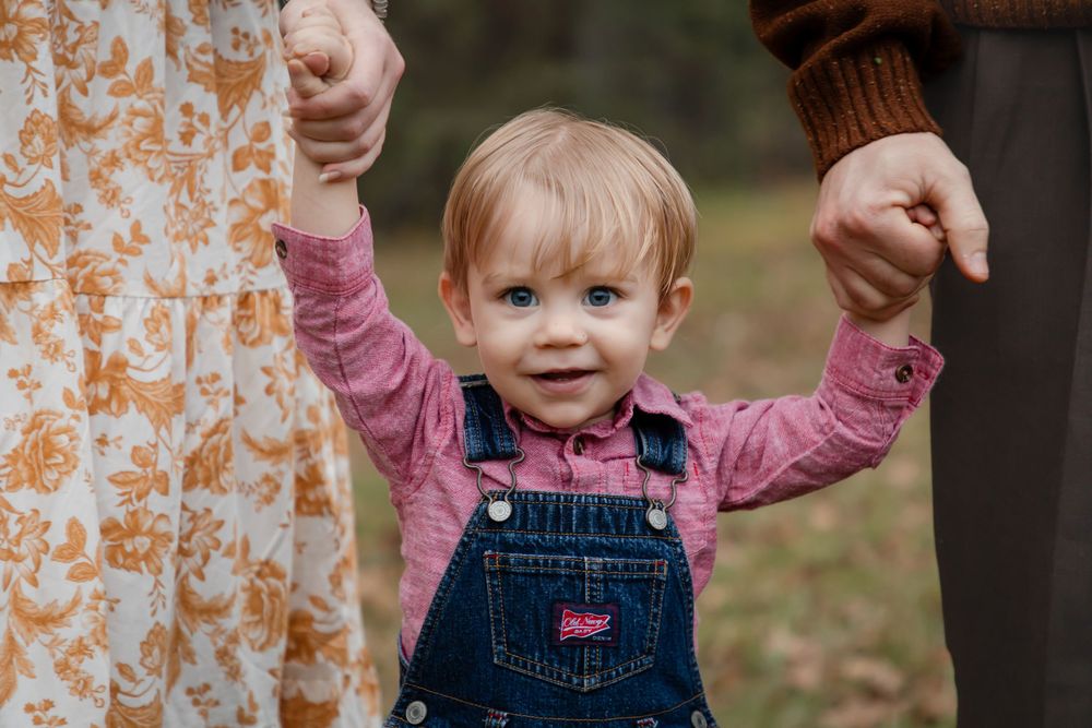 Small child wearing denim overalls and pink bow jumps joyfully in front of a floral curtain background.