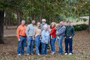 Multi-generational group poses together on fallen autumn leaves in a wooded outdoor setting.