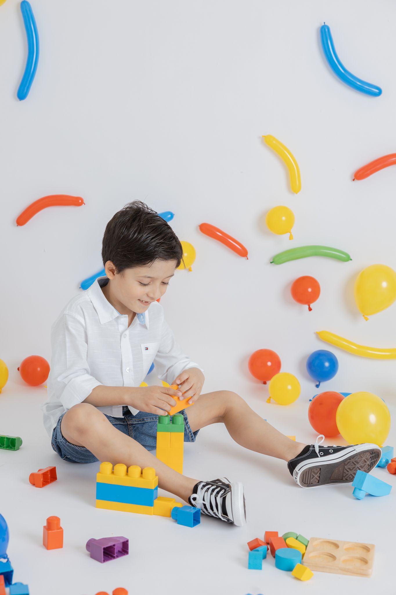 Child playing with colorful building blocks and toys on white background with balloon decorations.