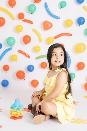 A child in a yellow dress poses playfully against a white backdrop decorated with colorful balloons and party decorations.
