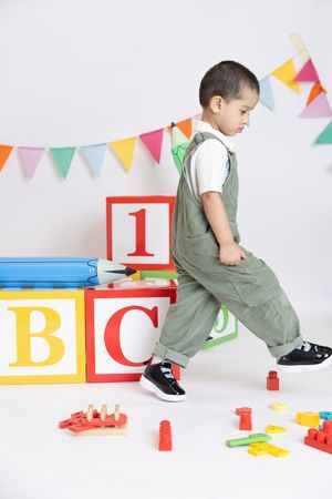 Child in olive green overalls plays with colorful alphabet blocks and toys under festive bunting decorations.