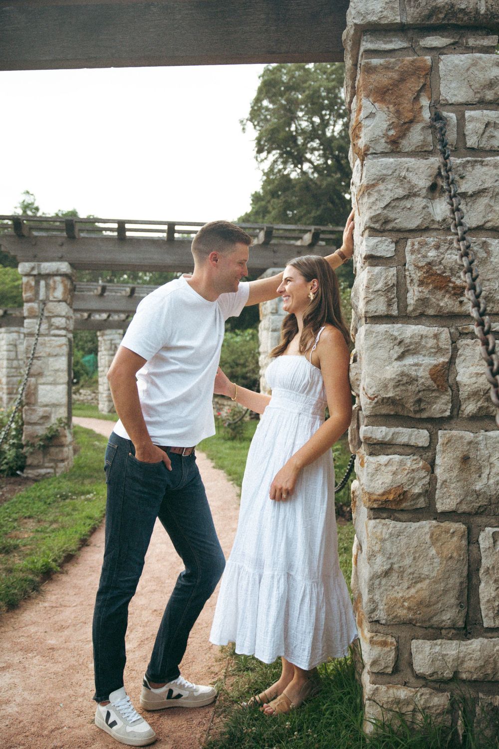 A romantic couple poses against a rustic stone structure with a wooden pergola in an outdoor garden setting.