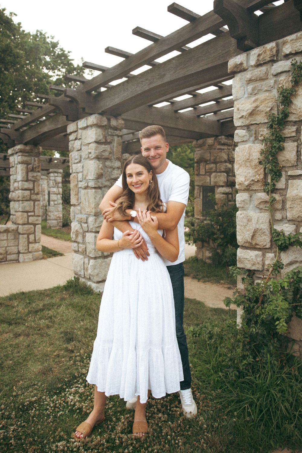 A couple embraces under a stone pergola structure covered in vines in a garden setting.