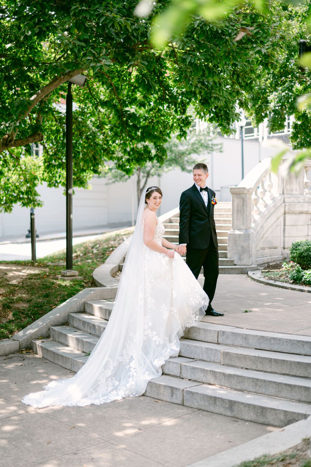 Wedding couple descends grand stone steps surrounded by lush green trees.