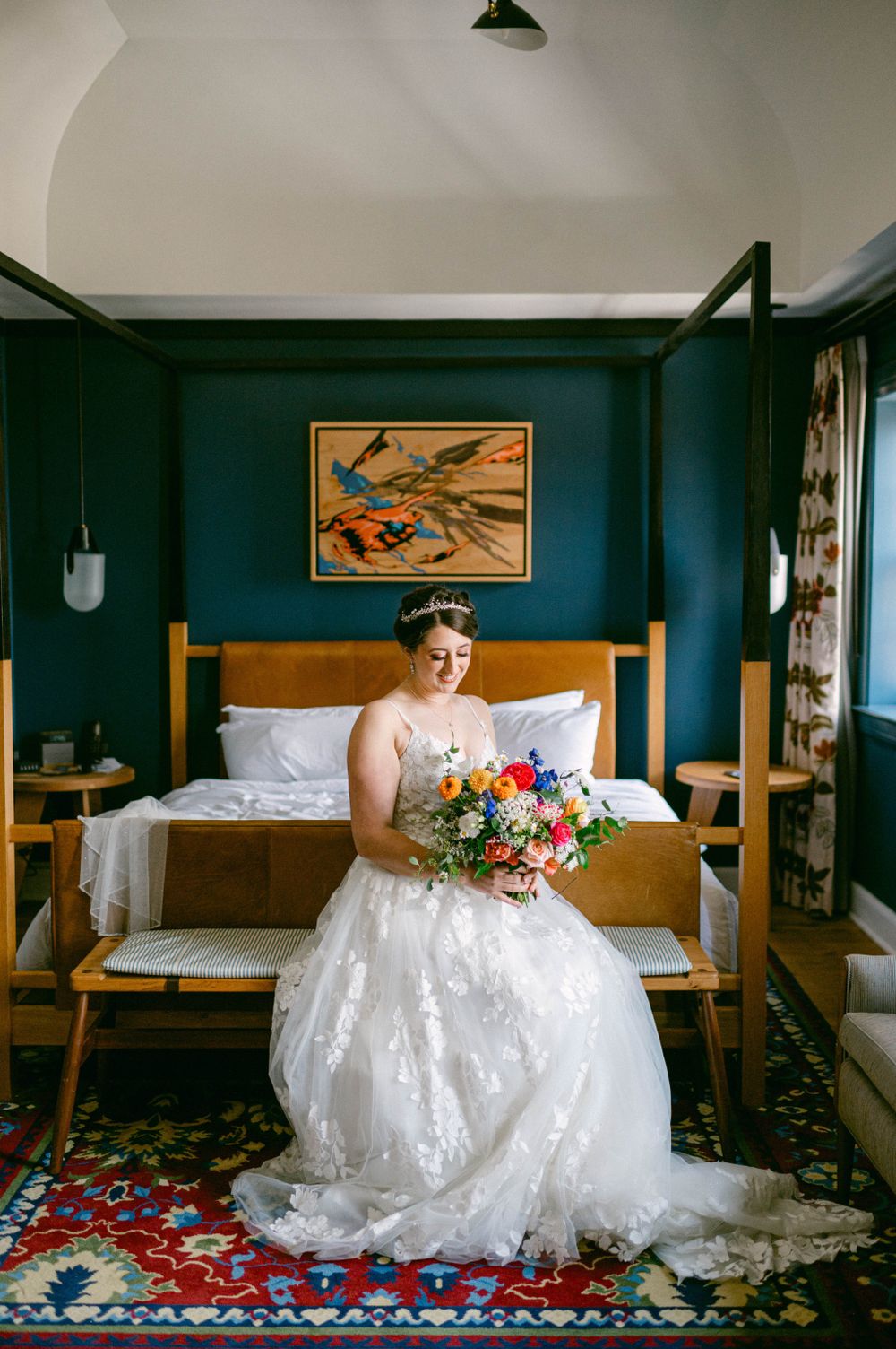 Bride in lace dress sits on hotel bed with colorful bouquet against deep teal walls.