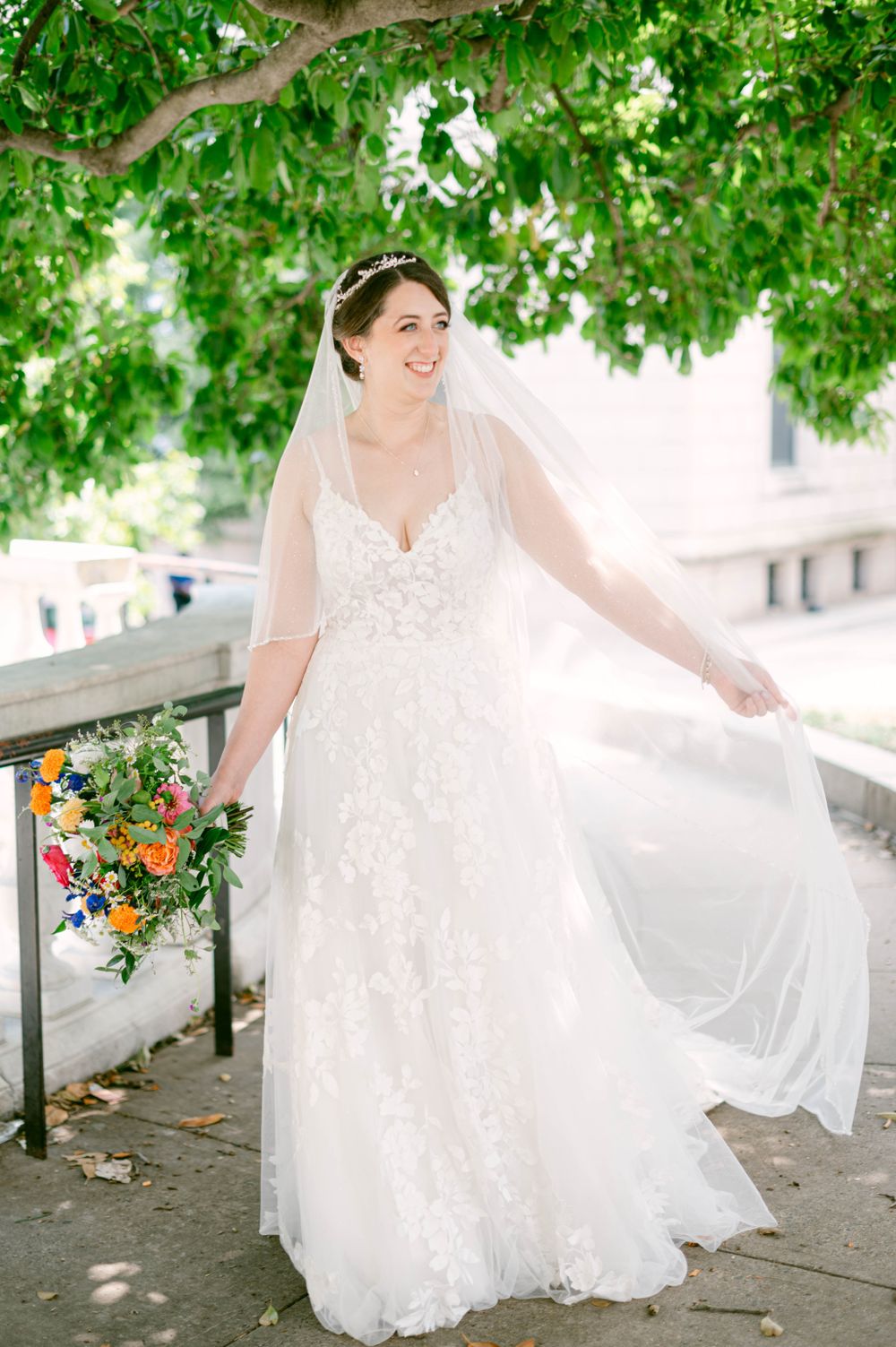 Bride in lace gown poses outdoors under green trees with colorful bouquet.