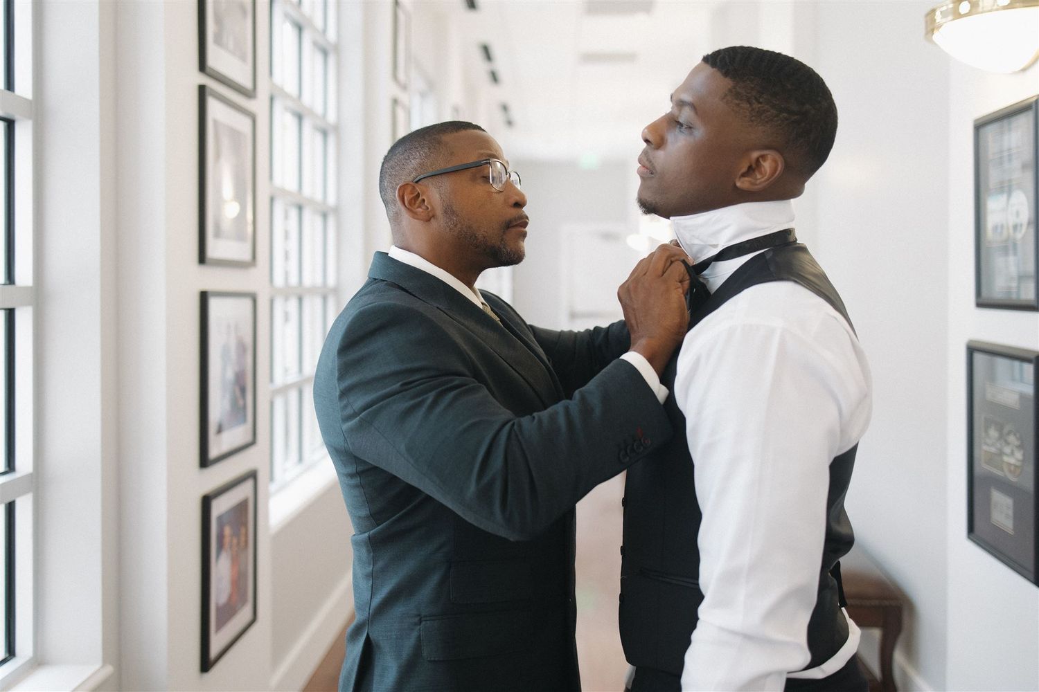 Two men in formal attire stand in a bright hallway while one adjusts the other's bow tie.