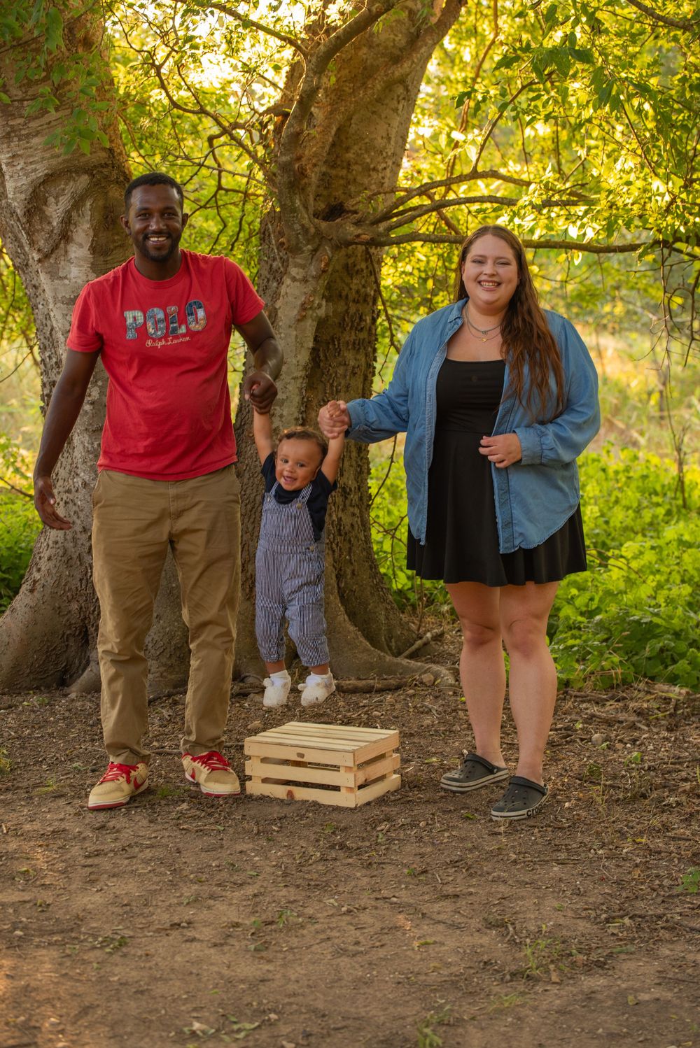 Family of three posing outdoors near a large tree, parents holding toddler's hands with a wooden crate nearby.