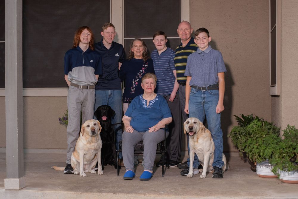 Family group photo with three Labrador dogs posing on a porch, featuring seven people of varying ages smiling together.