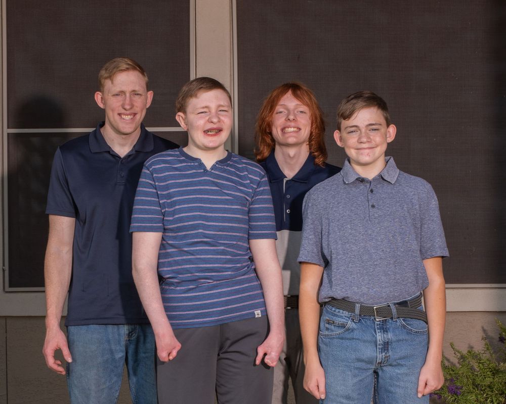 Four smiling boys posing together outdoors in front of a building, wearing casual polo and striped shirts.