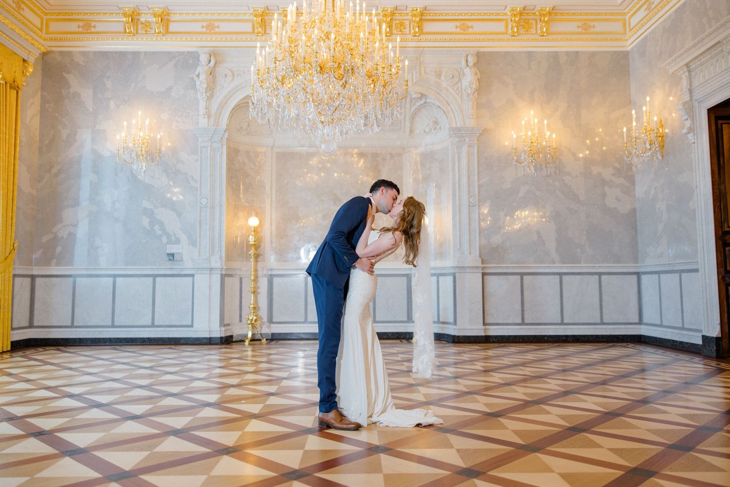 Two people dance in an elegant ballroom with ornate chandeliers and checkered marble floors.
