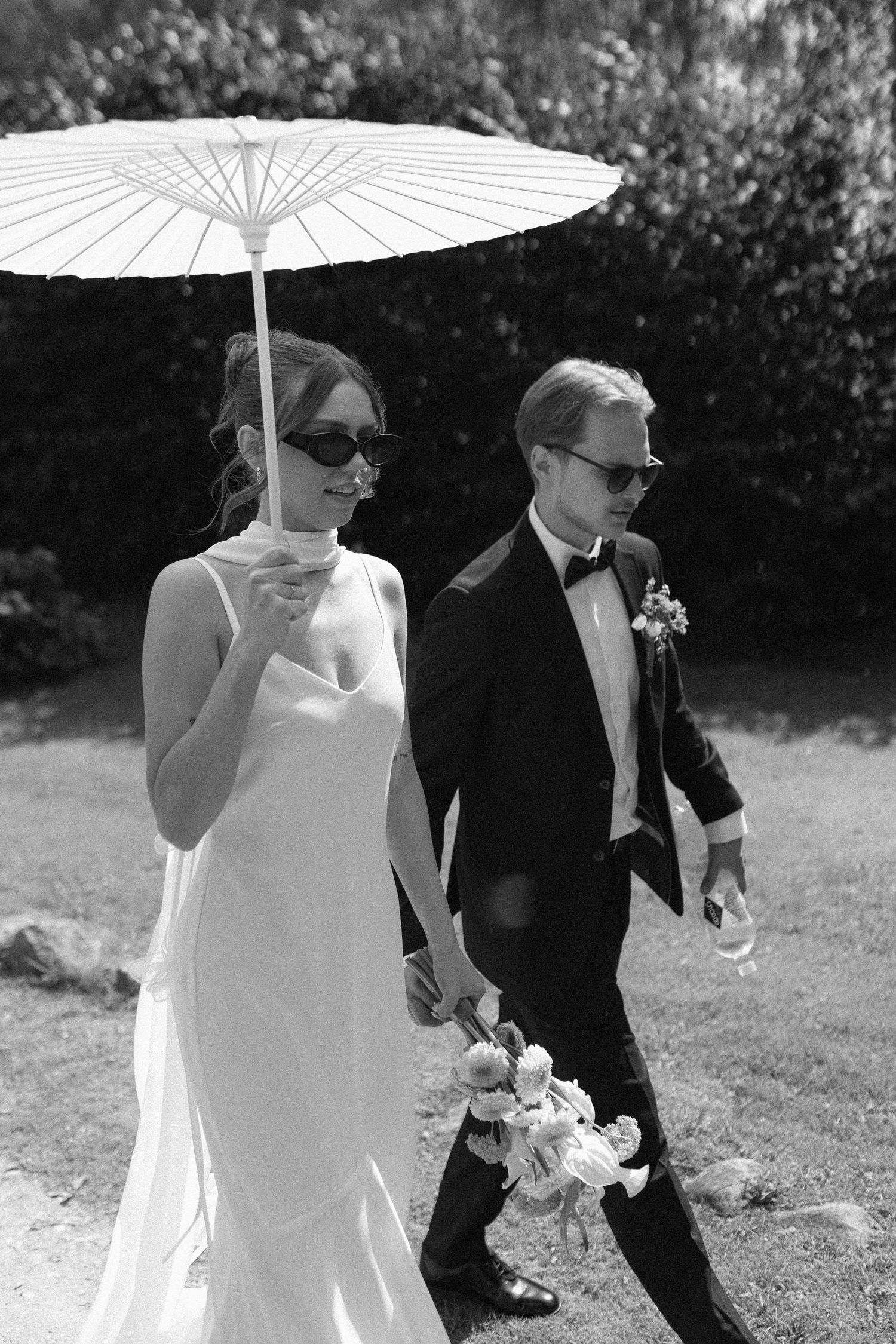 Wedding couple walks through garden holding white parasol umbrellas.