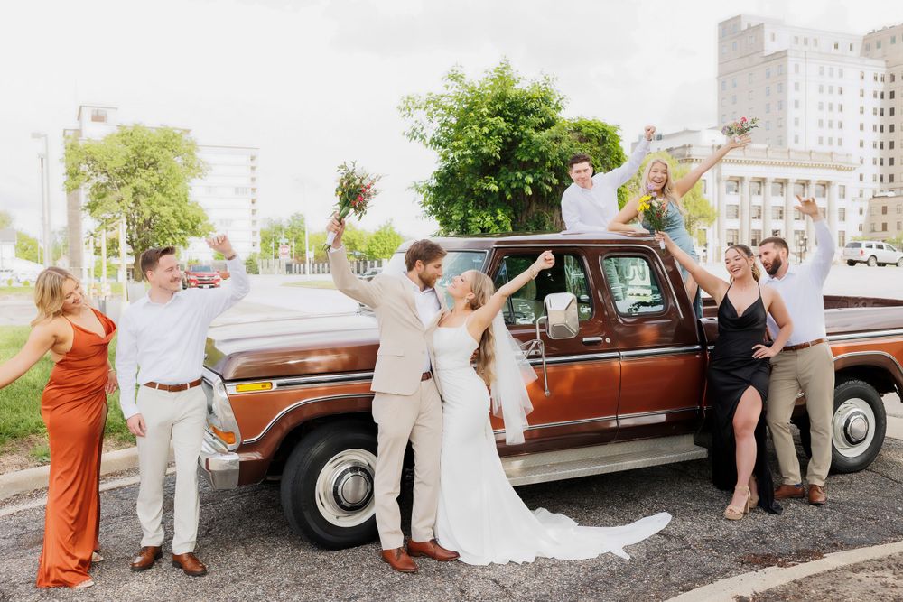 Wedding group celebrates beside classic brown Ford truck with city buildings in background.