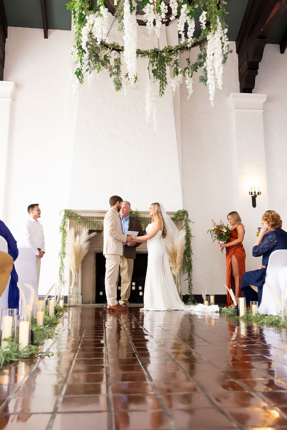 Wedding ceremony under a dramatic floral chandelier in a bright white room with terracotta flooring.