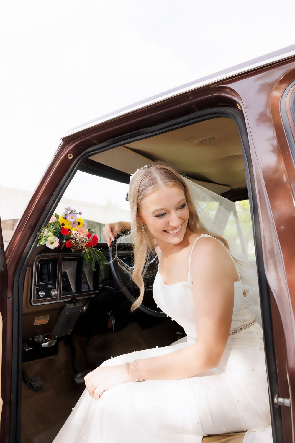 A bride in a white wedding dress sits in a vintage brown truck with colorful flowers visible.