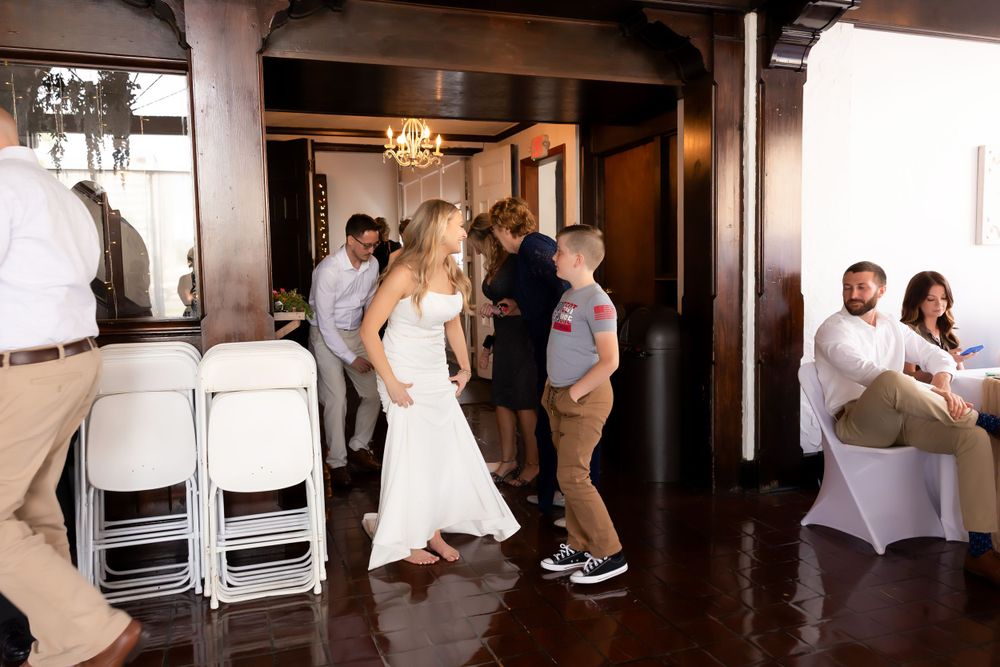Wedding party dances and celebrates in an indoor reception venue with stacked chairs visible.