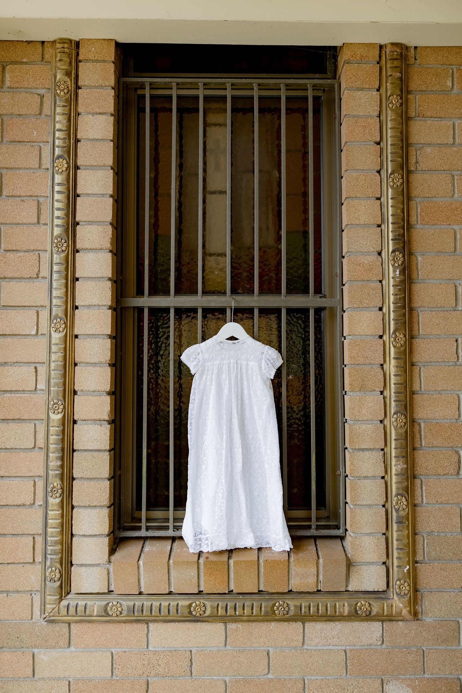 White christening gown hanging on barred window against brick wall.