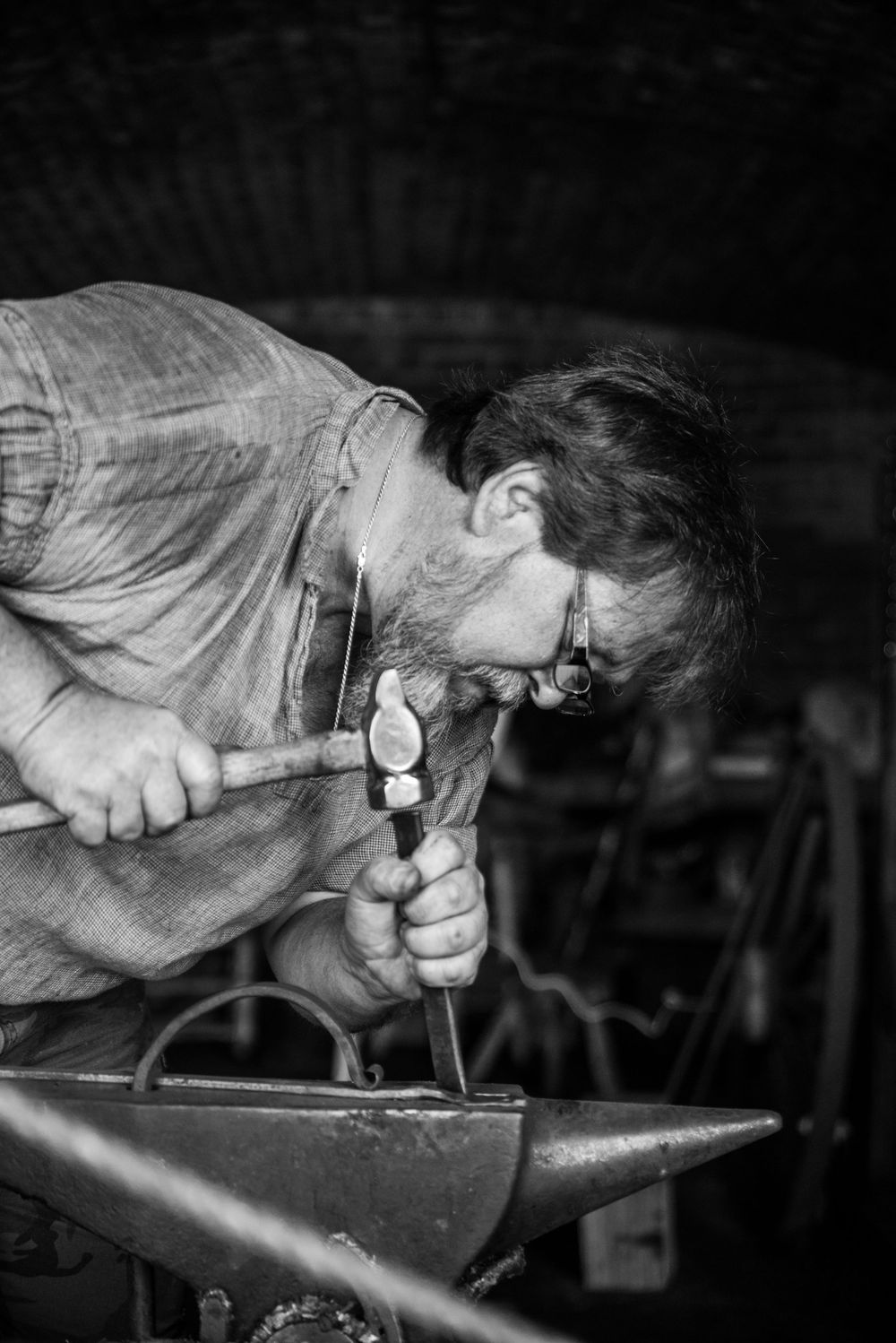 Craftsman in workshop intensely focused while hammering metal on an anvil in black and white photography.