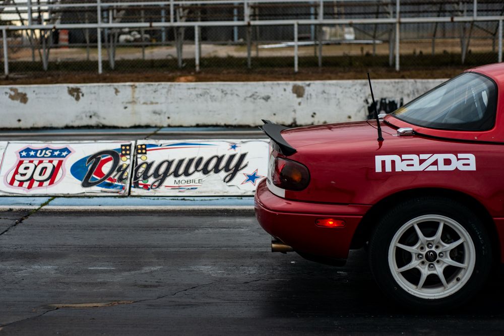 A red Mazda Miata sports car parked next to a white Lagaway racing barrier at a motorsports venue.