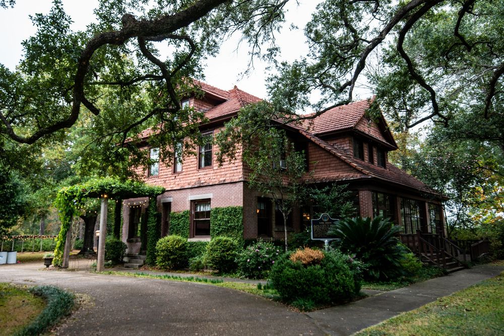 Historic Victorian mansion with red brick exterior and wraparound porch nestled among oak trees in a lush garden setting.