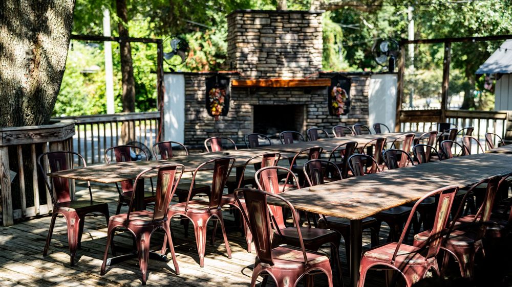 Rustic outdoor restaurant patio with wooden tables and chairs arranged under trees near a stone fireplace.