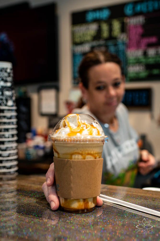 A caramel frappuccino topped with whipped cream and drizzle sits on a cafe counter with colorful decor in the background.