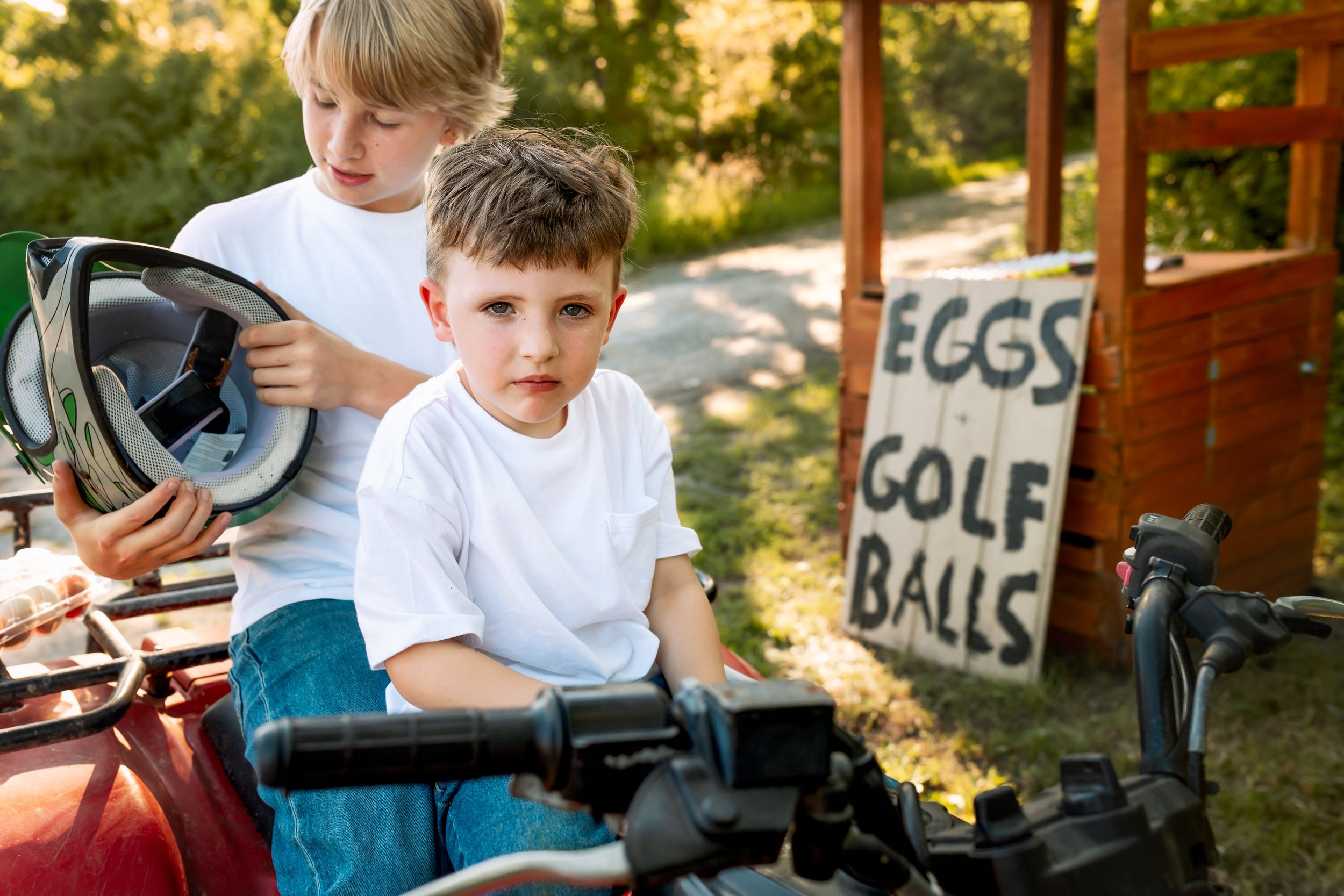 Two young boys in white shirts sit on a red ATV outdoors, one holding a helmet, beside a wooden sign reading eggs and golf balls.