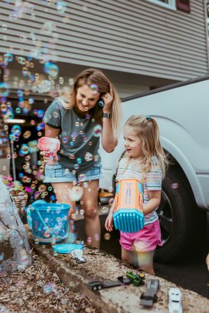 Two children playing with bubbles and colorful toys on a summer day outside a house with a car visible.