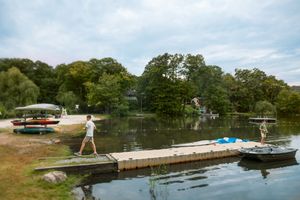 People walk along a wooden dock beside a calm lake, with kayaks stored on a rack and lush green trees lining the peaceful shoreline.