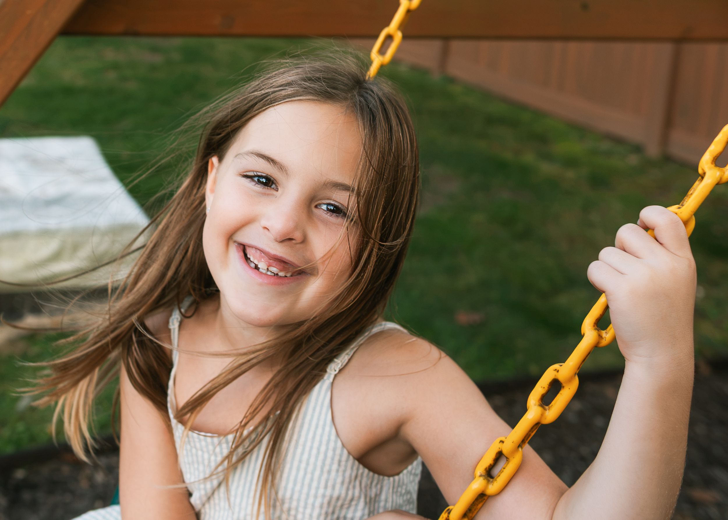 Smiling young girl with long brown hair sits on a playground swing holding a yellow chain in a sunny backyard.