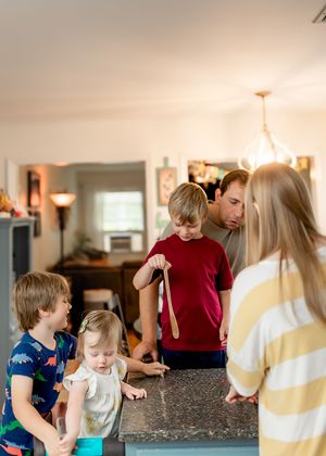 A family gathers around a kitchen counter to prepare food together in a warm, well-lit home setting.