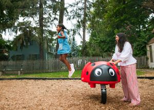 Young girl in a blue dress bouncing off a red ladybug spring rider as a woman in striped pants watches at a wooded playground.