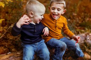 Two young children in casual sweaters laugh together while sitting outdoors during autumn.