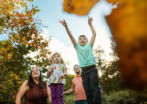 Happy family enjoying an autumn day outdoors as a boy in a teal shirt raises his arms while a girl, woman, and another child stand nearby.