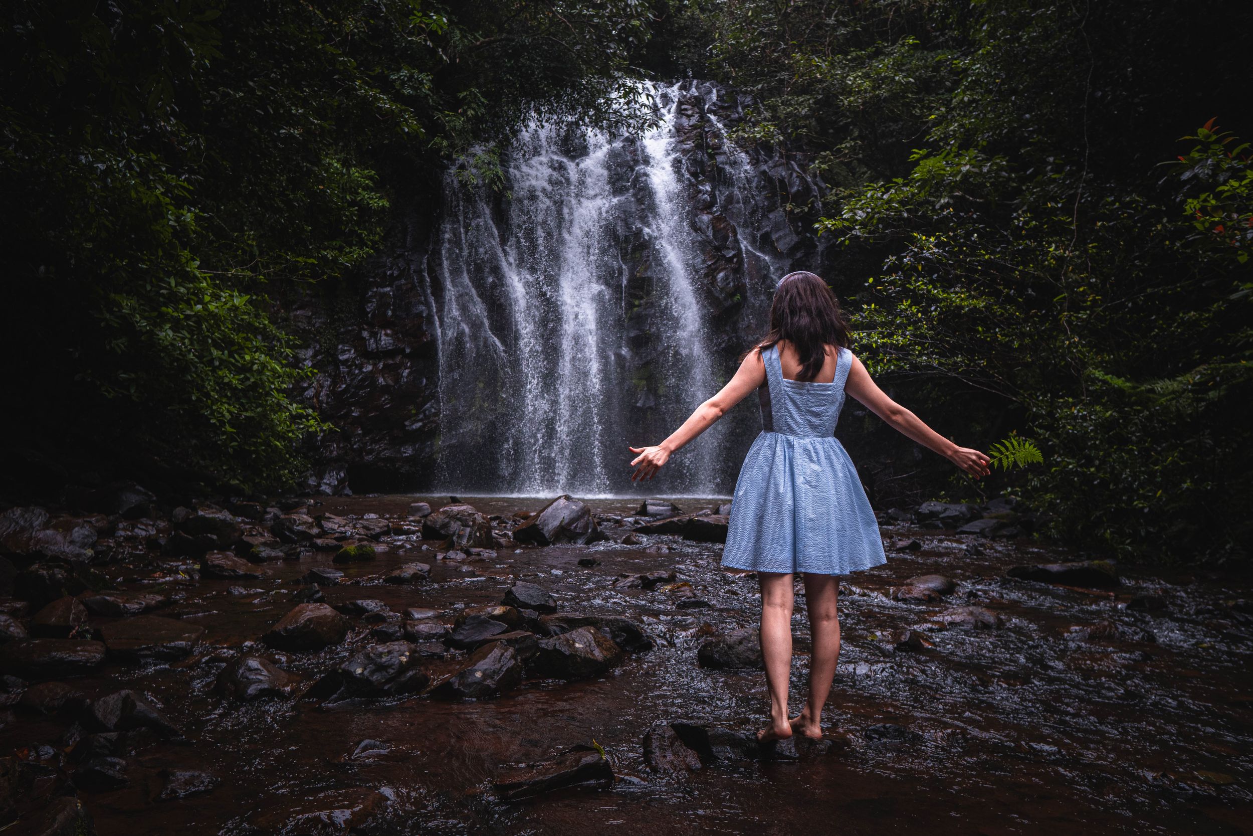 Asian Figure stands with outstretched arms before a cascading waterfall in the forest.